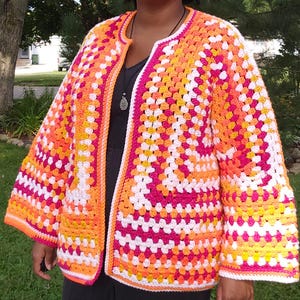 May include: A crocheted cardigan with a white background and colorful granny squares in shades of pink, orange, and yellow.