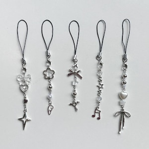 Puede incluir: Cinco correas de teléfono con dijes de plata con diferentes dijes, que incluyen una flor, un lazo, una nota musical, un corazón y un par de alas. Cada correa tiene un lazo de cuerda negro.