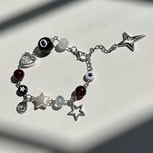 Puede incluir: Un brazalete de cadena plateada con cuentas negras y blancas, un dije de coraz&oacute;n, un dije de estrella y un dije de bola de billar.