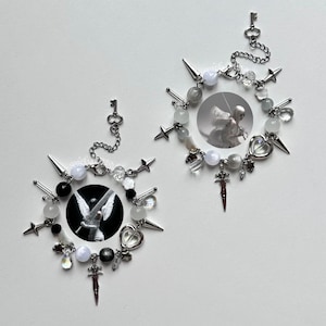 May include: Two circular charm bracelets featuring silver-tone chains, spikes, star charms, heart-shaped pendants, and sword charms. Each bracelet includes a central circular image of an angel figure and a small key charm attached to the chain.