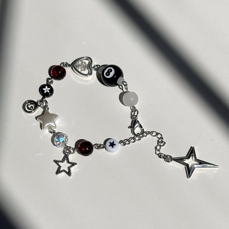 Puede incluir: Un brazalete de plata con un dije de bola de billar negra y blanca, cuentas rojas y blancas y dijes de estrellas plateadas.