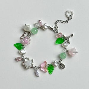 May include: A silver charm bracelet with a variety of charms, including flowers, leaves, hearts, and a key. The charms are made of glass and metal and are in shades of pink, green, and white.