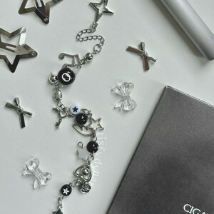May include: A silver charm bracelet with black and white beads, charms, and a star pendant. The bracelet has a music note charm, a key charm, a heart charm, and a star charm. The bracelet is on a white surface.