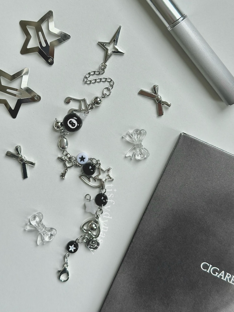 May include: A silver charm bracelet with black and white beads, charms, and a star pendant. The bracelet has a music note charm, a key charm, a heart charm, and a star charm. The bracelet is on a white surface.