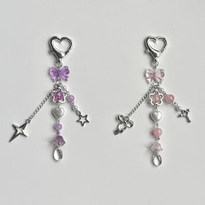 May include: Two silver keychains with charms. The keychain on the left has purple and white charms, including a star, a heart, a flower, and a bow. The keychain on the right has pink and white charms, including a key, a heart, a flower, and a bow.