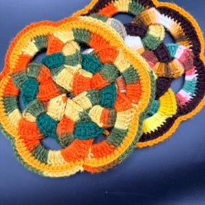 May include: Two handmade, colorful crochet trivets. The trivets are flower-shaped with a central hole. The color scheme includes orange, yellow, green, and brown. The trivets are made of yarn and are suitable for kitchen use.