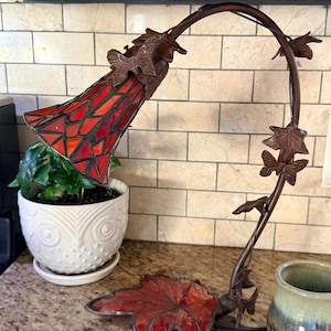 May include: A decorative table lamp with a stained glass shade in shades of red and orange. The lamp has a bronze-colored, curved stem with leaf and butterfly accents. The base of the lamp is a red leaf shape.