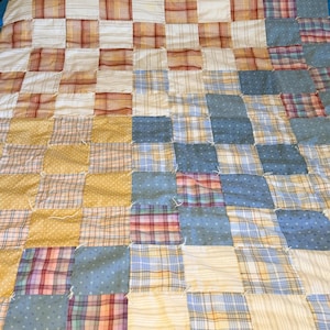 May include: A patchwork quilt with a grid pattern of squares in various colors and patterns. The quilt features squares of white, yellow, blue, and red plaid and striped fabrics. The quilt is on a bed with a blue border.