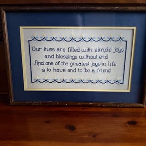May include: Framed cross-stitch artwork with a navy blue mat and a dark brown wooden frame. The cross-stitch features the text "Our lives are filled with simple joys and blessings without end. And one of the greatest joys in life is to have and to be a friend."