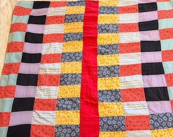 May include: A patchwork quilt with a vibrant mix of colors and patterns. The quilt features a central red stripe flanked by columns of yellow, blue floral, and red patterned blocks. Other blocks include black, lavender, and light blue stripes. The quilt is a handmade textile.