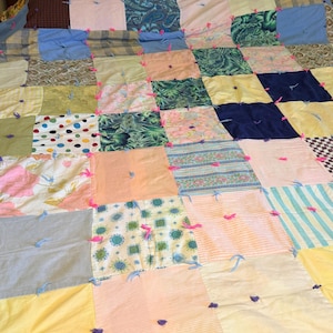 May include: A patchwork quilt with a variety of fabric squares in pastel colors, including pink, blue, yellow, and green. The quilt features a mix of patterns, such as stripes, polka dots, and floral prints, and is adorned with pink and blue thread accents.