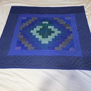 May include: A square quilt featuring a pixelated diamond pattern in shades of blue, teal, and grey. The center displays a geometric motif surrounded by concentric borders of varying blue tones, all finished with a diamond-shaped quilted texture.