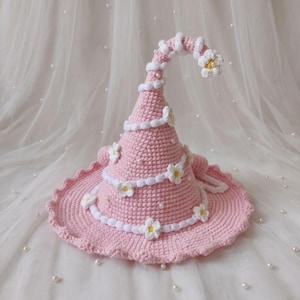 May include: A light pink crocheted pointed hat featuring a spiraling white yarn trim, small white flower appliqués with yellow centers, and scattered white pearl beads. The hat has a wide, slightly ruffled brim and a curved, tapered top section.