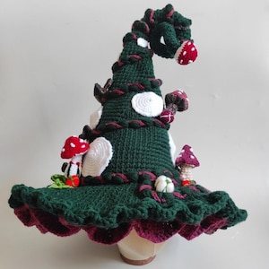May include: A dark green crocheted hat with a tall, curled point, featuring attached three-dimensional crocheted mushrooms in red and white, white circular patches, and a wide, ruffled brim with a deep burgundy underside and dark green trim.