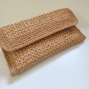 Könnte beinhalten: Eine rechteckige Clutch aus hellbraunem, geflochtenem Strohmaterial. Die Tasche weist eine texturierte, ineinandergreifende Struktur auf der gesamten Oberfläche auf, einschließlich eines umgeklappten Verschlusses am oberen Teil.