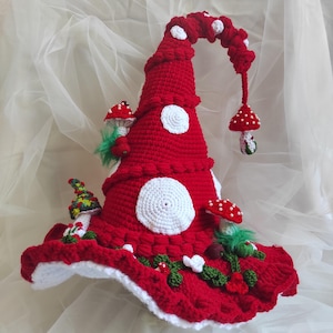 May include: A red crocheted hat with a tall, pointed, drooping top. The hat is decorated with white circular patches, several small crocheted mushrooms with red caps and white spots, green yarn accents, and a dangling mushroom charm.