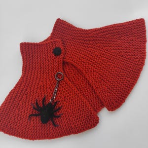May include: A bright red knitted cowl scarf featuring a textured garter stitch pattern. The accessory includes a small black button closure at the top and a dangling black spider charm attached to the fabric by a thin silver-toned metal chain.
