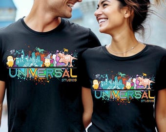May include: Black t-shirts with a vibrant graphic featuring the words "UNIVERSAL STUDIOS" and cartoon characters. The design includes a cityscape. A smiling couple is wearing the shirts.