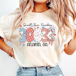 May include: Cream-coloured t-shirt with the text "Small Town Freedom" and the numbers "30033" in a patriotic design. The text "ATLANTA, GA" is printed below the numbers. The shirt features red, white, and blue accents.
