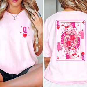 Puede incluir: Camiseta rosa con un gr&aacute;fico de una carta de reina de corazones en la parte posterior y un peque&ntilde;o gr&aacute;fico de reina de corazones en la parte delantera.