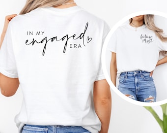 May include: White t-shirt with black text that reads "In my engaged era" with a small heart symbol. The shirt is being worn by a person with long brown hair. A second white t-shirt with black text that reads "Future Mrs." with a small circle symbol is shown in the background.
