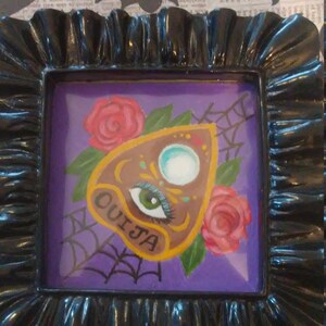 May include: A black framed picture with a purple background and a painted image of a brown Ouija board with a green eye and a white circle in the centre. The board is surrounded by two pink roses and a spider web.