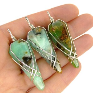 May include: Three green gemstone pendants wrapped in silver wire. The gemstones are a light green with brown and black inclusions. The pendants are all different shapes and sizes.
