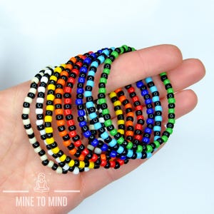May include: A collection of colorful beaded bracelets in a variety of colors, including black, white, yellow, orange, red, green, and blue. The bracelets are held in a hand against a white background. The bracelets are made of small, round beads.