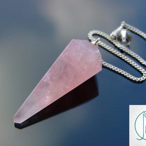 Könnte beinhalten: Ein Rosenquarz-Pendel mit einer Silberkette. Das Pendel hat eine hellrosa Farbe und eine spitz zulaufende, dreieckige Form. Die Kette ist oben am Pendel befestigt und besteht aus kleinen, verbundenen Metallstücken. Der Hintergrund ist eine dunkle, reflektierende Oberfläche.