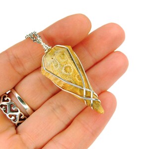 May include: A silver wire wrapped pendant featuring a fossilized coral stone with a light brown and yellow color. The stone has a unique pattern of circular shapes.
