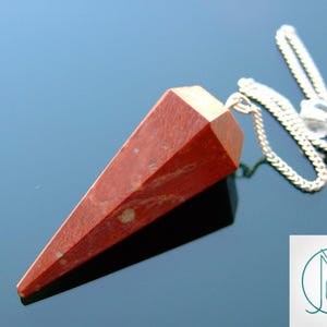 May include: Red jasper pendulum with a silver chain. The pendulum is a pointed, faceted crystal with a reddish-brown colour and white flecks.