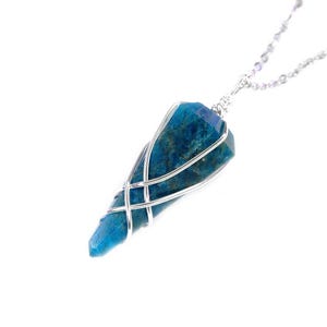 May include: A blue gemstone pendant necklace with a silver chain. The gemstone is a deep teal color, encased in a silver wire cage. The pendant is a long, pointed shape. The necklace is displayed against a white background.
