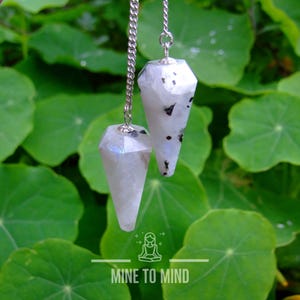 May include: Two white rainbow moonstone crystal pendulums with black flecks hanging from silver chains against a green leafy background. The text "MINE TO MIND" is at the bottom of the image.