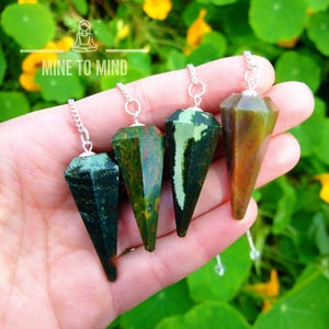 May include: Four green, brown, and white gemstone pendulums with silver chains. The pendulums are held in a hand against a green and yellow background.  Mine to Mind.