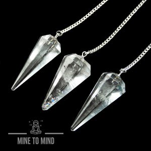 May include: Three clear quartz crystal pendulums with silver chains. The pendulums are hanging against a black background.