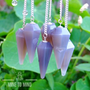 May include: Six gray and purple crystal pendulums with silver chains hang against a backdrop of green leaves. The pendulums have a faceted, geometric design and a pointed bottom. The image includes the text "MINE TO MIND".