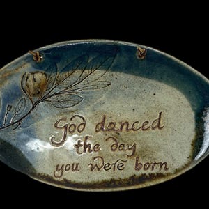 May include: Oval ceramic plaque with a blue and beige glaze. The plaque features an embossed floral design and the inscription "God danced the day you were born" in a brown color. Two holes are at the top for hanging.
