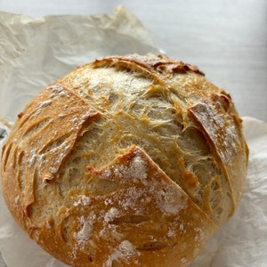 Peut inclure: Une miche de pain au levain ronde avec une croûte dorée et croustillante, marquée d'une incision en forme de croix sur le dessus. La surface est légèrement saupoudrée de farine blanche, révélant une texture alvéolée dans les entailles.