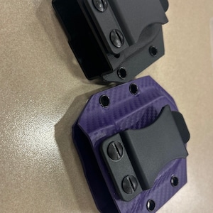 May include: Two gun holsters, one black and one purple with a carbon fiber pattern. Both holsters have a black clip.