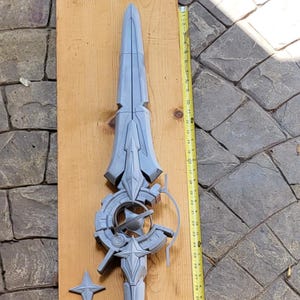 May include: A light gray, unpainted 3D-printed spear prop featuring an intricate, mechanical-style head with circular and star-shaped elements. A yellow measuring tape placed alongside indicates the total length is approximately 70 inches.