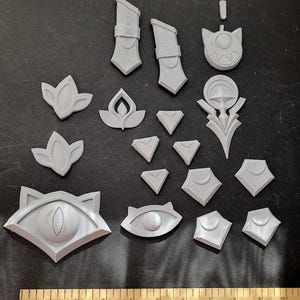 May include: A collection of gray, 3D-printed decorative pieces, including stylized eye shapes, geometric forms, and floral designs. A wooden ruler with inch markings is at the bottom of the image.