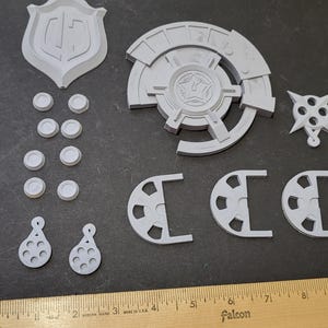 May include: A collection of gray 3D-printed components, including a shield shape, circular elements, and other geometric forms. The pieces are arranged on a dark surface, with a wooden ruler providing scale.