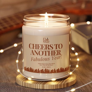 May include: A clear glass candle jar with a white soy wax candle inside. The candle has a brown label with the text "CHEERS TO ANOTHER Fabulous Year!" and the words "100% Natural Soy Wax Blend, Burn Time 50-60 Hours, 9 OZ/255 G".