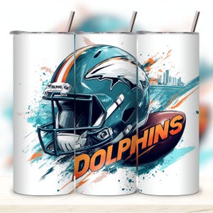 May include: A white tumbler with a teal, orange, and white Miami Dolphins football helmet graphic. The helmet has a dolphin logo and the word "Dolphins" is written in orange on a brown football.