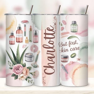 May include: A white tumbler with a stainless steel straw, featuring a watercolor design of skincare products, aloe vera, and roses. The text "Charlotte" is displayed vertically, and "But first, skin care" is also present.