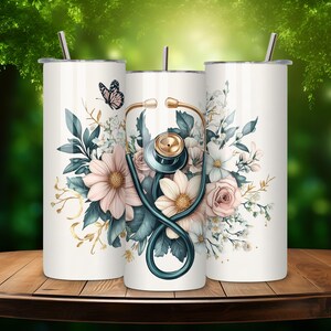 Puede incluir: Tres vasos de acero inoxidable con fondo blanco y un dise&ntilde;o floral con un estetoscopio y una mariposa. Los vasos est&aacute;n sobre una mesa de madera.