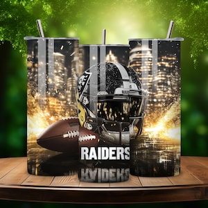 K&ouml;nnte beinhalten: Drei Edelstahlbecher mit einem schwarz-wei&szlig;en Design, das einen Footballhelm mit dem Wort "RAIDERS" zeigt. Der Hintergrund ist ein verschwommenes Bild einer Skyline.