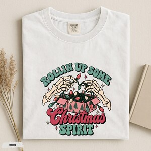 May include: A white t-shirt featuring a graphic of two skeleton hands reaching into a pink box filled with colorful Christmas lights. The text reads "ROLLIN UP SOME Christmas SPIRIT" in a retro font. The shirt has a Comfort Colors brand label.