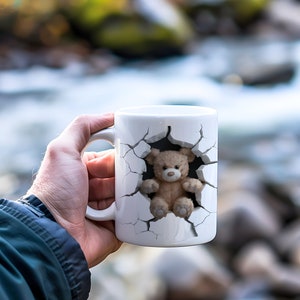 Könnte beinhalten: Weiße Keramiktasse mit einer 3D-Illustration eines Teddybären, der aus einer zerbrochenen Wand kommt. Der Teddybär ist hellbraun mit einer dunkelbraunen Nase und Knopfaugen. Die Tasse hat einen weißen Henkel.