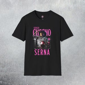May include: Black t-shirt featuring a pink graphic of Sergio Checho Serna. The graphic includes a photo of a person and the name "Sergio Checho Serna" in pink text. The shirt is a crew neck.